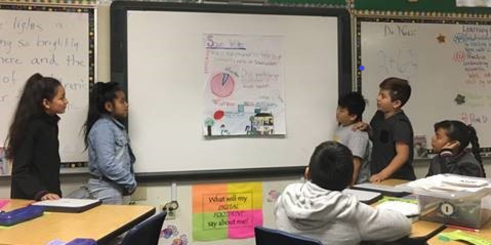 four students stand at a hand-drawn poster while other students sit at classroom desks