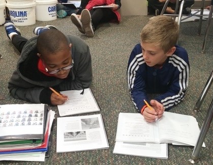 Two students sit on the floor of their classroom and write down information in notebooks.