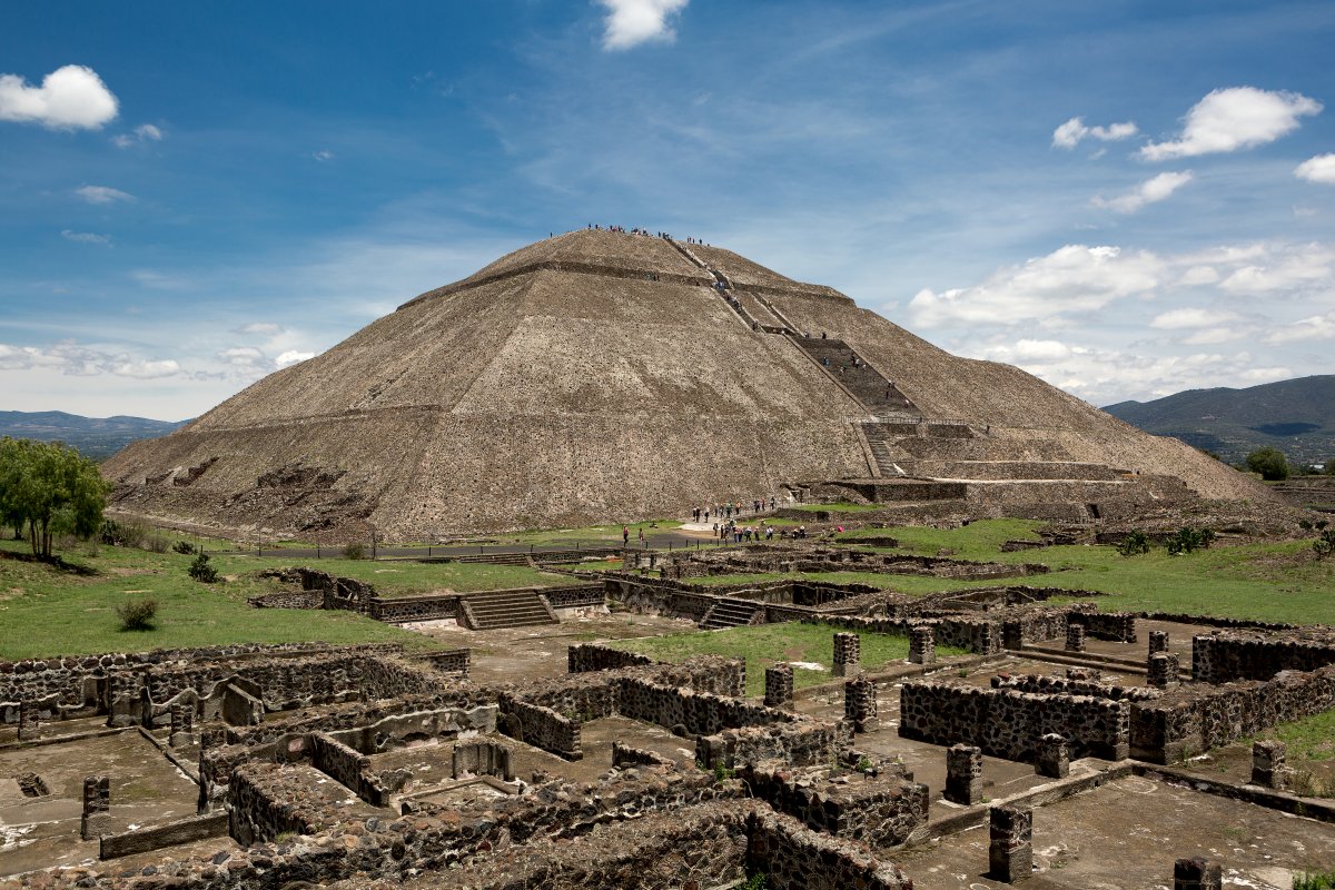 A wide pyramid with people walking up its steps and standing on the flat top and civilization ruins at the base of the pyramid