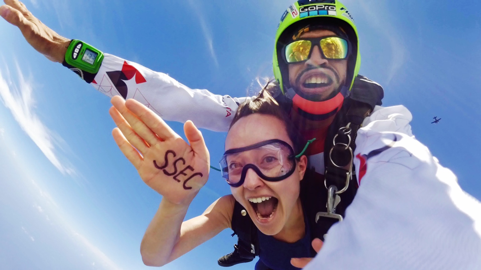 Skydiver in free-fall, hand outstretched. SSEC is written on her hand. Male instructor behind.